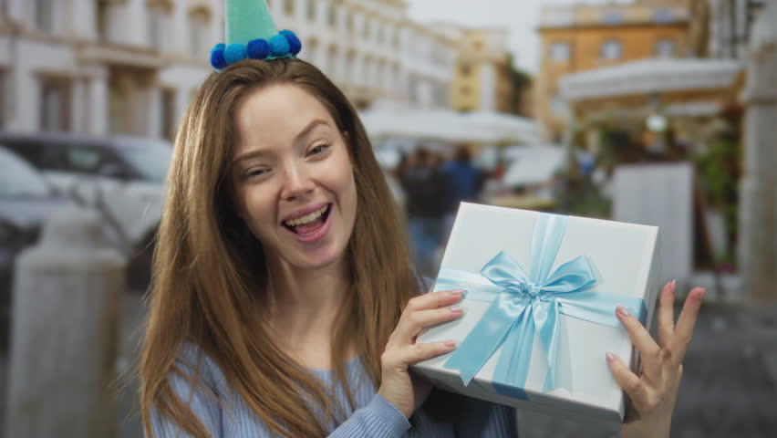 Woman wearing party hat smiling and holding gift box with blue ribbon on street; happiness celebration.