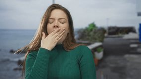 Woman covers mouth with hand on a seaside street lined with planters and coastal rocks over blue ocean waves; tiredness. - Powered by Shutterstock - Get 15% off with code: PIKWIZARD15