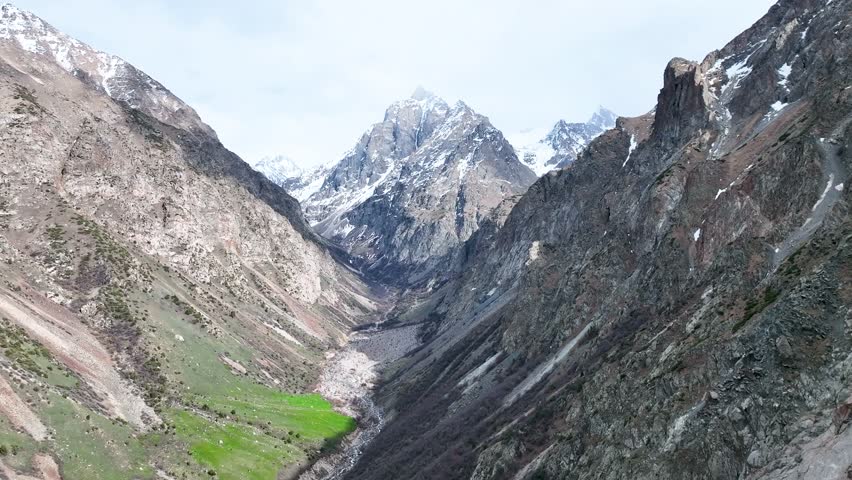 Dramatic view deep into a rugged mountain gorge with sheer rock walls, a stream flowing through the valley floor, and a snow-capped peak in the distance