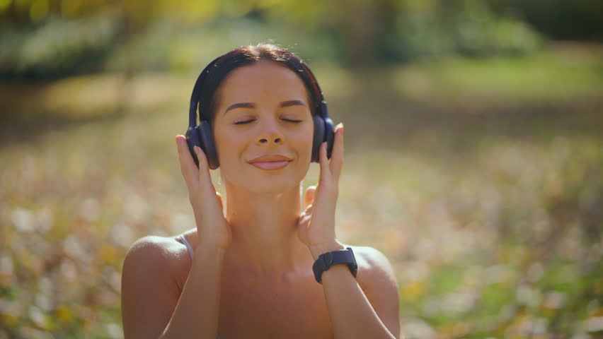 Cheerful young woman enjoying her favorite music in wireless headphones while relaxing in a sunlit park, smiling peacefully with her eyes closed in a moment of pure bliss and calm