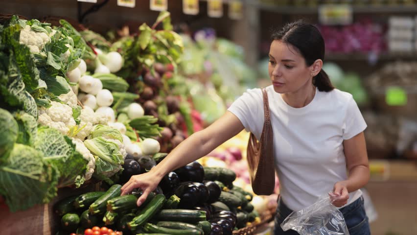 Young woman shopping in vegetable store and buy zucchini. Customer carefully examine goods, thinking about buying. Self-service store, vegetables and fruits from all over world
