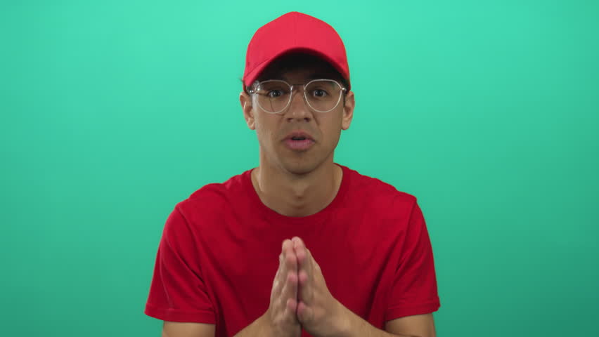Young hispanic man in red shirt and cap holds hands pressed in prayer gesture in green studio; hope.