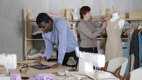 Focused experienced fashion designer working on digital sketch of new garment using tablet in studio while female colleague working on fitting dress on mannequin in background - Powered by Shutterstock - Get 15% off with code: PIKWIZARD15