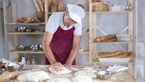 Experienced mature baker in maroon apron kneading pliable dough on floured work surface in cozy artisan bakery against backdrop of wooden shelves lined with bread loaves, baguettes and pastries - Powered by Shutterstock - Get 15% off with code: PIKWIZARD15