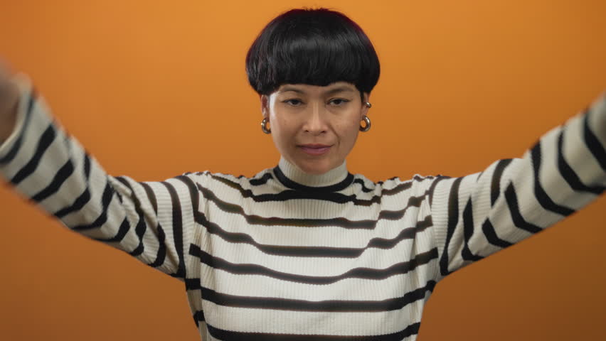 Young chinese woman making horn gestures with both hands on head against an isolated orange background, wearing a striped sweater and smiling playfully.