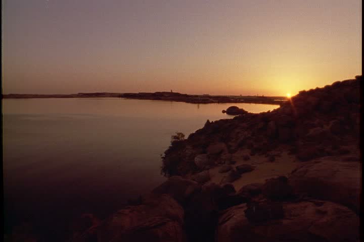WS of sun setting behind rocky island. Camera pans left across Lake Nasser in Aswan, Egypt.