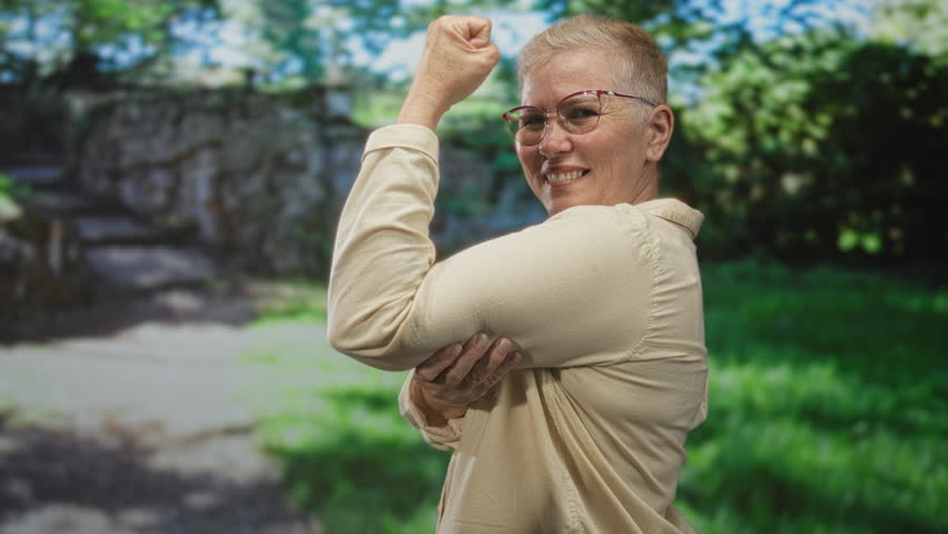 Woman flexes arm showing biceps and smiles while standing near a stone arch in a sunlit forest with green foliage and stone steps; confidence.