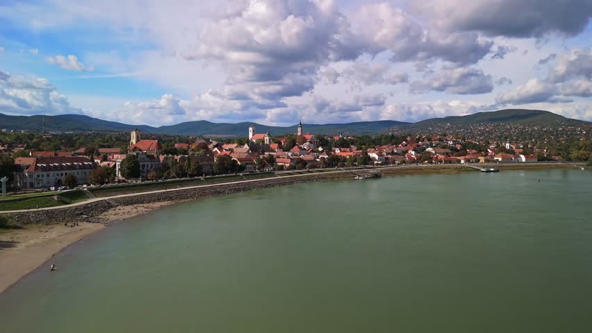 Low-level drone flight above the Danube River toward the colorful town of Szentendre, Hungary, with its red rooftops, historic churches, and scenic hills under a partly cloudy sky.
