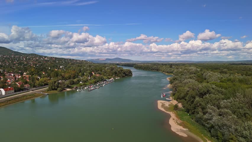 Drone flying forward north over the Danube River at the Szentendre suburb, Hungary, showcasing lush riverbanks, small marinas, and scenic hills under a bright sky filled with clouds.