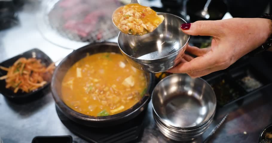 Close-up shot of a steaming earthenware pot of Doenjang Jjigae Korean stew being scooped with a ladle into a silver metal bowl next to side dishes