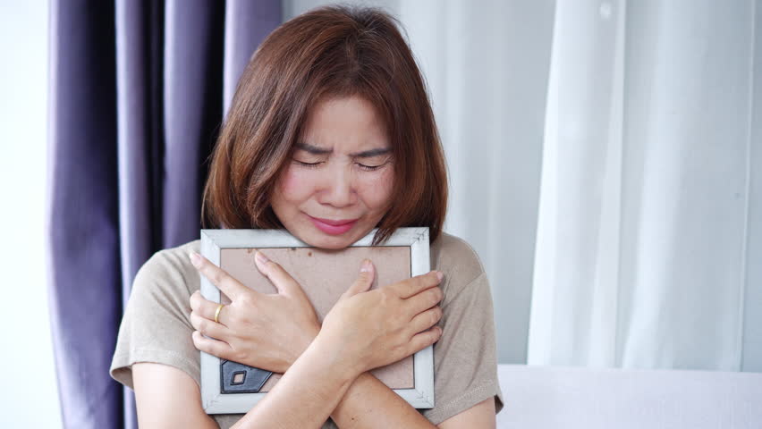 Grieving Asian woman crying while holding a photo of a loved one who passed away, expressing deep sadness, loneliness, and emotional pain. Concept of mourning, loss, heartbreak, and emotional healing