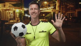 Man referee holding soccer ball with palm raised on street in neon yellow jersey and whistle visible; fair play authority. - Powered by Shutterstock - Get 15% off with code: PIKWIZARD15
