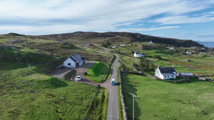 Aerial footage of a blue camper van driving through a village on the North Coast 500. Scenic Scottish Highlands landscape with white cottages, rolling hills, and ocean views under blue skies.