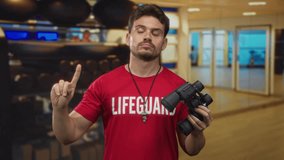 Man lifeguard in red shirt with whistle points finger upward in gymnasium while holding binoculars near exercise balls and mirrored wall; vigilant duty. - Powered by Shutterstock - Get 15% off with code: PIKWIZARD15