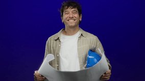 Man smiling holding blueprints with hardhat against a vibrant blue background, showcasing confident hispanic professional demeanor and stylish casual attire. - Powered by Shutterstock - Get 15% off with code: PIKWIZARD15
