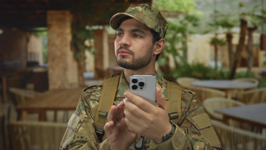 Young hispanic man in camouflage military uniform holds smartphone with both hands and looks upward on street; duty.