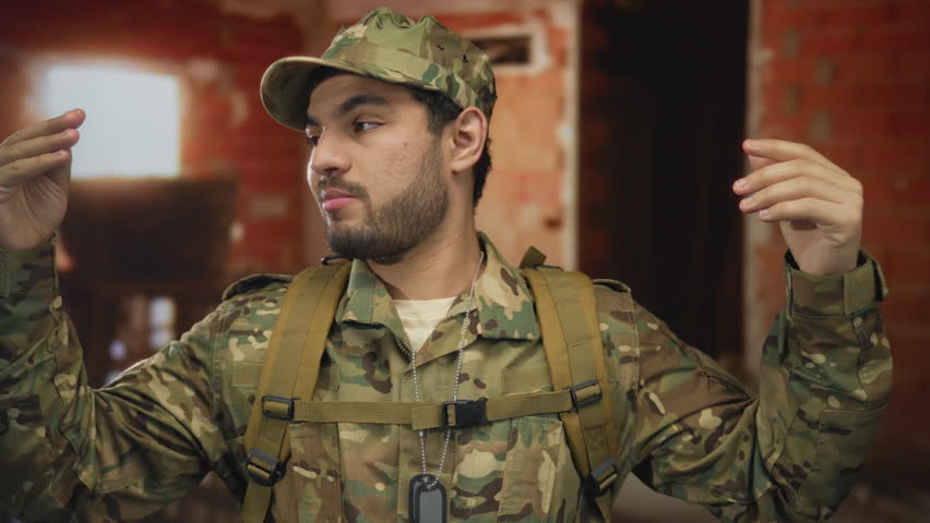 Man in camouflage uniform with backpack and dog tags raises both hands inside a brick building; duty focus discipline readiness.