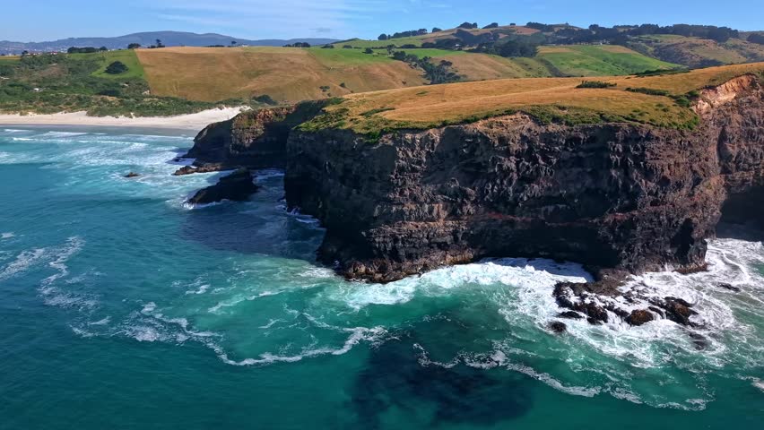 Drone side shot moving left to right along the cliffs of Smaills Beach, showing waves crashing on rocks, green vegetation, and a blue sky.