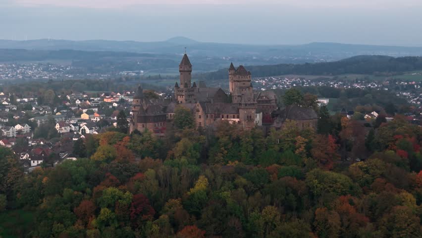 Slow aerial retreat from medieval castle on forested hilltop with autumn foliage overlooking German town under moody sunset sky