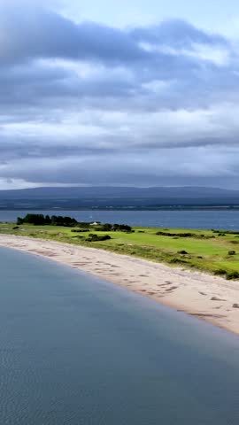 Drone glides above sandy shoreline, grassy fields, and water under dramatic cloudy summer sky