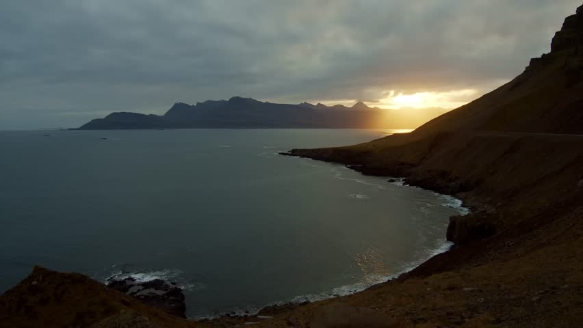 A view of the rugged coastline and dramatic mountains of the East Fjords region in Iceland. The footage captures the serene beauty of the landscape at sunset, with the low sun casting a warm glow over