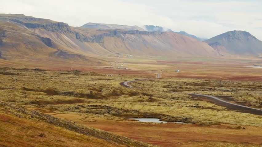 A wide angle shot of a vast and beautiful landscape in Iceland, featuring a winding road cutting through a golden hued, moss covered plain towards a dramatic mountain range under a cloudy sky.