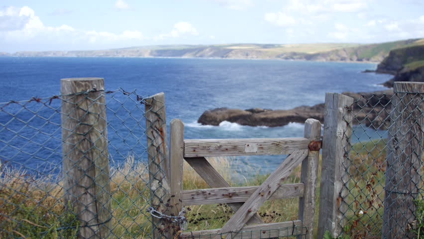 A fence with a wooden gate is standing at the edge of a coastal path above the blue waters near Port Isaac and Portwenn, capturing Cornwall