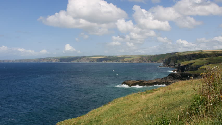 Cliffs overlooking the ocean are stretching along the rugged coastline of Cornwall near Port Isaac and Portwenn, with waves crashing below and rolling hills in the distance