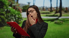 Hispanic woman in glasses holding a red clipboard, yawning outdoors in a sunny park, suggesting fatigue or boredom, with lush greenery in the background. - Powered by Shutterstock - Get 15% off with code: PIKWIZARD15