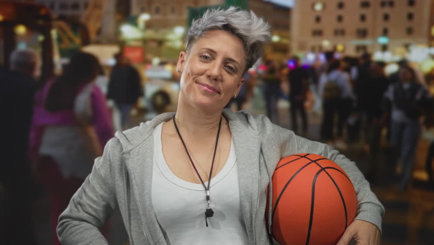 Woman coach with short hair holding orange basketball on bustling city street; confidence determination empowerment.