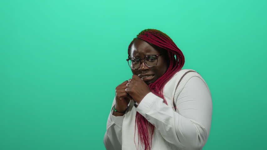 Woman with pink braids and glasses poses nervously against an isolated green background wall wearing a white shirt expressing emotion vividly highlighting modern urban style.
