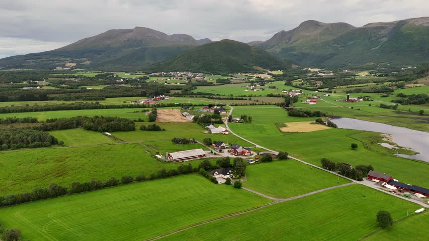 An aerial view of lush farmland in Fraena, Norway, with red farmhouses, green fields, and low, distant mountains beneath an overcast sky