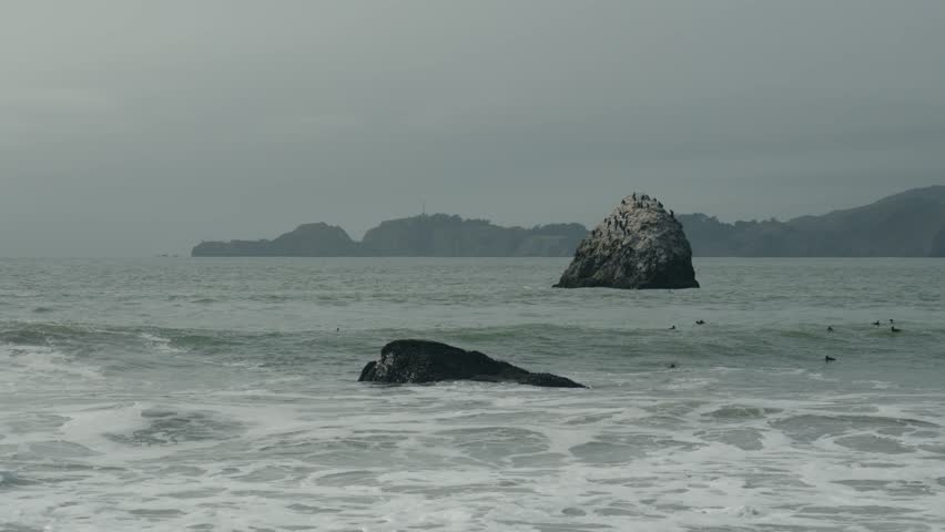 A wide coastal view of waves crashing against large rocks in a calm gray sea, with distant hills visible beneath an overcast sky