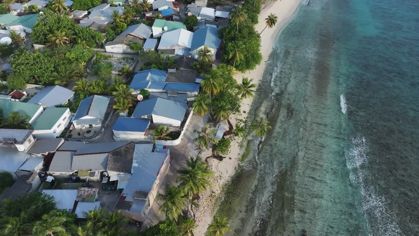 Aerial view of a coastal village with houses nestled among lush trees along a white sandy beach meeting the turquoise waters, Maalhos, Baa Atoll, Maldives.