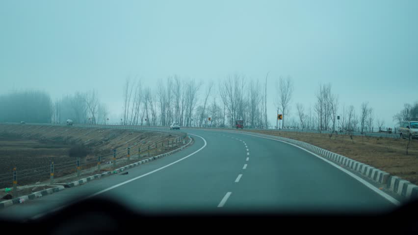 4K shot of mountain road in foggy winter morning at Kashmir, India. Dry trees and heavy fog on sides. POV shot of car driving on dreamy mountain road. Roadtrip and travel concept.