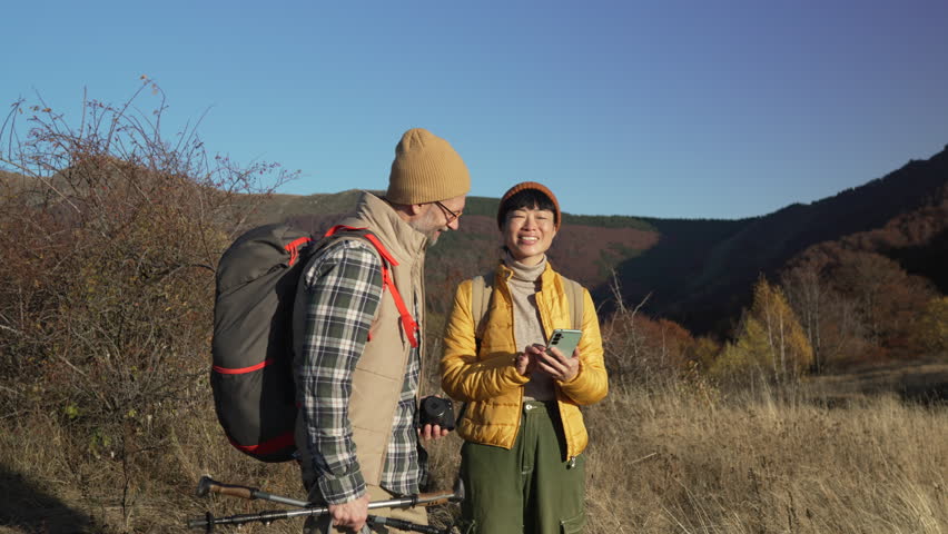 Middle-aged interracial couple stopping during a mountain hike, the woman using a smart phone for navigation while pointing towards the path, enjoying the beautiful sunny autumn landscape