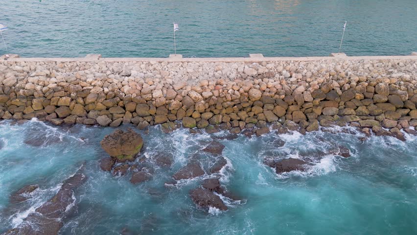 Aerial view of waves crashing against the rugged stone barrier in Old Jaffa, contrasting the turquoise water with the weathered rocks, Tel Aviv-Yafo, Israel.