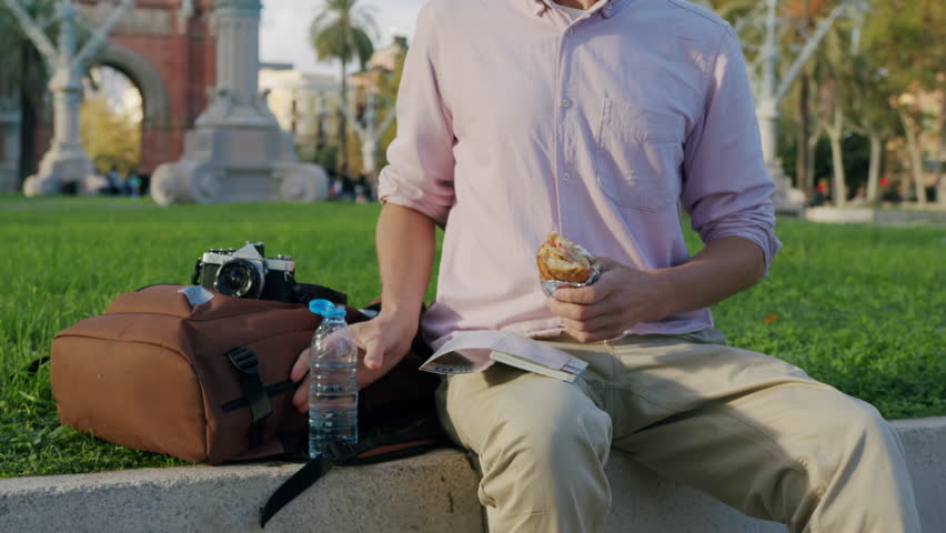 Young man office worker having lunch outdoors, drinking coffee and eating sandwich while sitting on bench near office building in park, tourist travel vacation in barcelona spain.