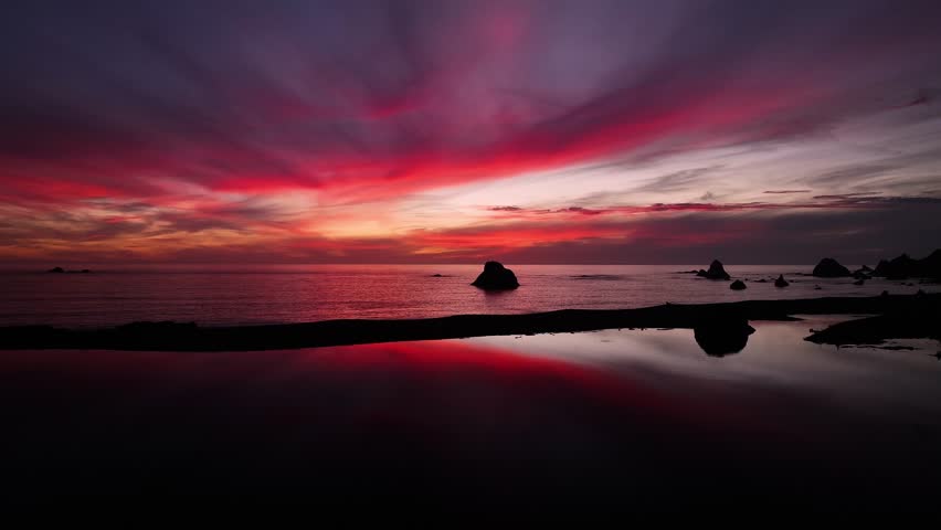 Aerial view of the Pacific ocean meeting the dark sandy beach, with rock formations silhouetted against a vibrant red and purple sunset, California, United States.