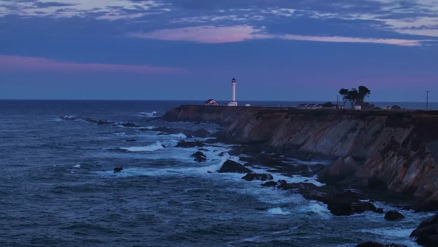 Aerial view of the Pigeon Point Lighthouse standing tall against the twilight sky, waves crashing against the rugged coastline, California, United States.