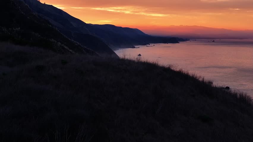 Aerial view of the rugged coastline with the sun setting, casting a golden glow on the water and silhouetting the mountains, California, United States.