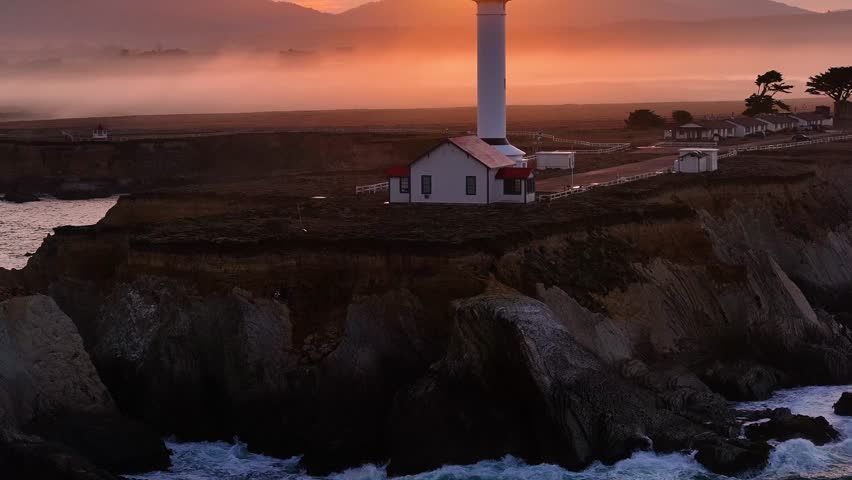 Aerial view of Point Arena Light stands tall against a fiery sunset, with fog adding a mystical touch to the landscape, Point Arena, California, United States.