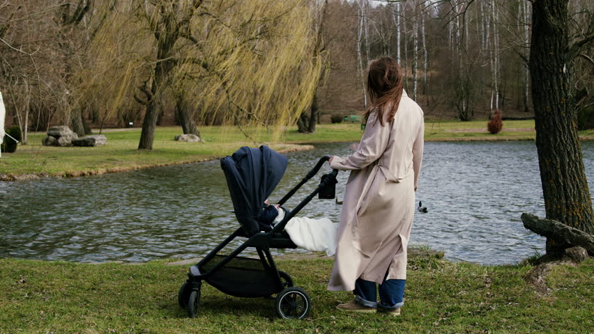 Woman in beige coat gently pushes stroller by serene lake, showcasing tranquil outdoor scene, camera follows her movement with smooth dolly motion