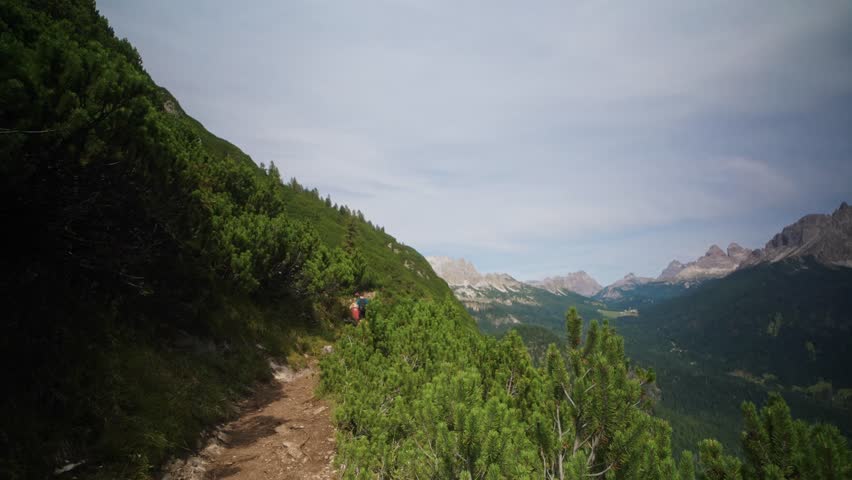 Hiker wearing a yellow jacket walking on a mountain trail near Lago di Sorapis in the Dolomites, Italy. Forest path with alpine peaks and summer landscape in the background