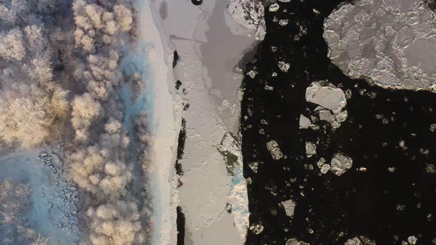 An aerial shot of a partially frozen river in the South Urals region of Russia. The footage captures the contrast between the dark, open water and the large, cracked ice floes. The shoreline is visibl
