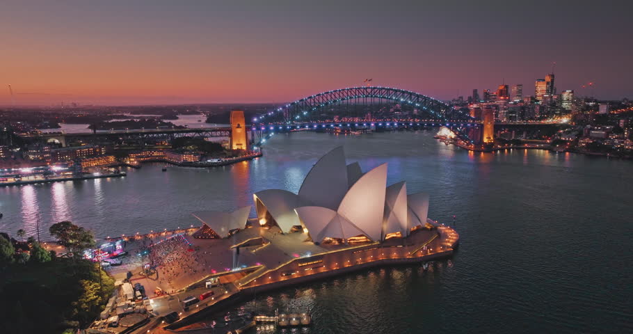 Australia, Sydney: aerial view of Sydney Opera House and Harbour Bridge illuminated at sunset, vibrant colorful evening sky over modern cityscape and harbor. Travel destination. Drone flight footage