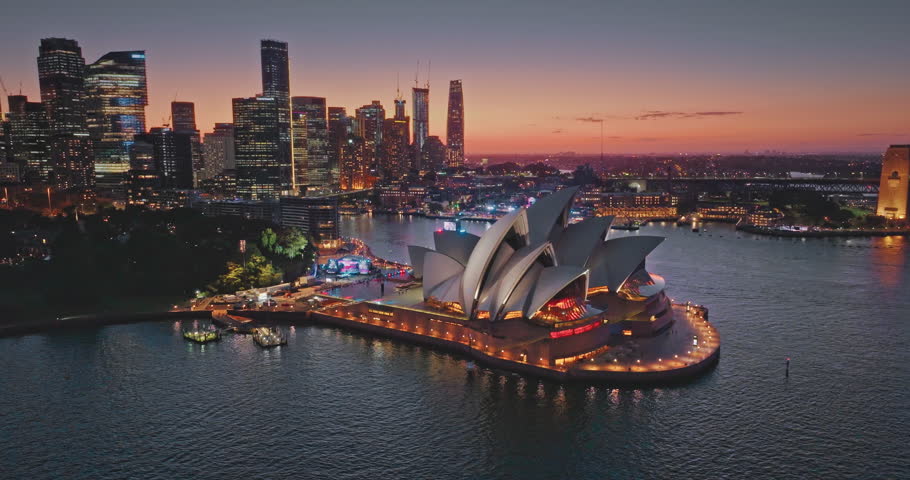 Australia, Sydney: Sydney Opera House glowing at colorful sunset with modern city skyline and illuminated buildings skyscrapers reflecting on Sydney Harbour. Travel destination. Drone flight footage