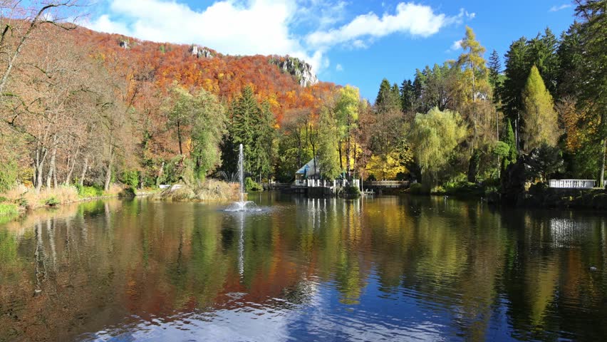 A serene lake reflects the vibrant fall foliage of the surrounding forest, complete with a charming fountain and clear blue skies above. Spa town Rajecke Teplice in Slovakia, Europe.