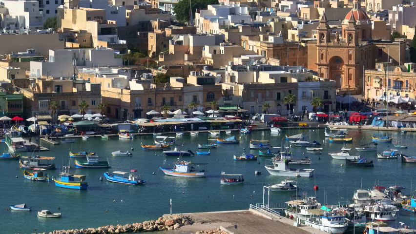 Malta, Marsaxlokk Harbour: Colorful fishing boats floating in Marsaxlokk Harbour, Malta, create a vibrant scene alongside a bustling market with vendors under umbrellas on a sunny day