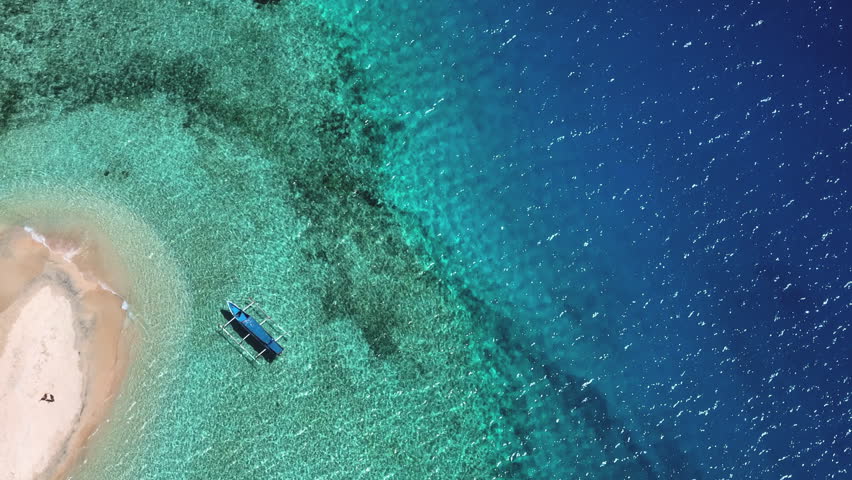 Aerial drone view of a traditional boat mooring near a small sandy beach and shallow coral reef, showcasing tropical vacation and pristine nature of the Gili Islands